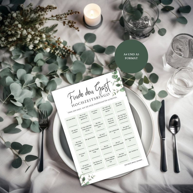 May include: A green and white wedding bingo card with the title "Finde den Gast Hochzeitsbingo". The card has 25 squares with different prompts to find a guest who matches the description. The card is on a white table with green leaves and a candle.