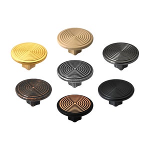 May include: Seven cabinet knobs in different metallic finishes. Each knob has a circular, concentric pattern on the face. Colours include gold, bronze, black, and silver. The knobs have a geometric base.