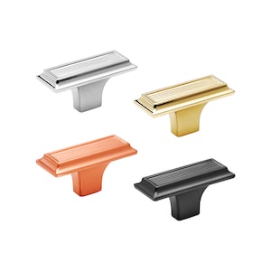 May include: Four cabinet knobs in silver, gold, copper, and black. Each knob has a rectangular, ridged top and a curved base. The knobs are designed for furniture and home decor.