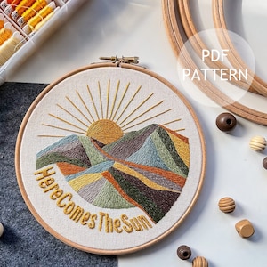 May include: A white embroidery hoop featuring a colourful embroidered design of a sun rising over mountains. The text "Here Comes The Sun" is embroidered below the mountains. The hoop is surrounded by wooden embroidery hoops, wooden beads, and a container of embroidery floss.