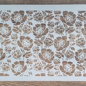 Könnte beinhalten: Weiße Kunststoffschablone mit einem floralen Design mit einem sich wiederholenden Muster aus Pfingstrosen. Die Schablone ist für Bastel- und Dekorationsarbeiten konzipiert.