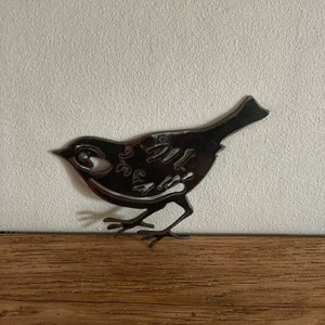 May include: A dark metal bird silhouette with intricate cut-out details. The bird has a round eye, a beak, and delicate legs. It is mounted on a wooden surface against a white wall. The bird is approximately 15 cm long.