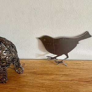 May include: A metal silhouette of a bird with a rustic finish. The bird is standing on a wooden surface.