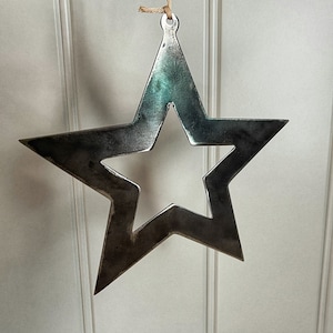 May include: A large, silver metal star ornament with a smaller star cut out in the centre. The star has a rustic, distressed finish and is hanging from a twine string.