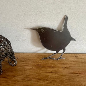 May include: A metal silhouette of a wren, painted black, with a rustic finish. The wren is standing on a wooden surface.