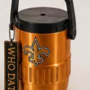 May include: A metallic gold insulated beverage container with a black lid and handle. The front features a fleur-de-lis emblem in gold, black, and white. A rectangular black tag hangs from the handle, displaying the text WHO DAT in gold letters.