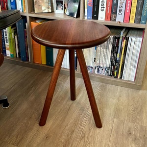 Könnte beinhalten: Ein dunkelbrauner Holz-Beistelltisch mit drei Beinen. Der Tisch ist rund und hat eine glatte Oberfläche. Der Tisch steht vor einem Bücherregal voller Bücher.