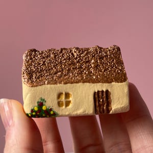 May include: A miniature, hand-painted cottage figurine. The roof is textured and brown, contrasting with the cream-colored walls. A small window and door are visible. Decorative flowers are painted at the base of the cottage.