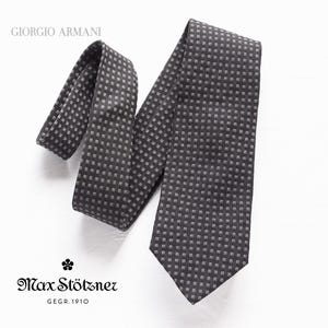 Può includere: Una cravatta di seta grigia scura con un motivo geometrico bianco. La cravatta è piegata e mostrata su uno sfondo bianco. Il testo "Giorgio Armani" è visibile nell'angolo in alto a sinistra dell'immagine.
