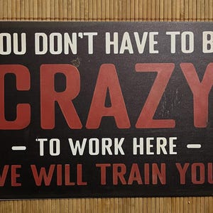 May include: Black metal sign with the text "YOU DON'T HAVE TO BE CRAZY - TO WORK HERE - WE WILL TRAIN YOU!" in white and red lettering. The sign has rounded corners and holes for hanging.
