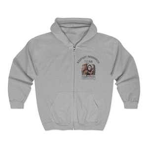 May include: A light gray zip-up hoodie with a Bigfoot Research Team graphic. The graphic features a Bigfoot illustration and the text "Hide and Seek Champion". The hoodie has a hood and a front pocket.