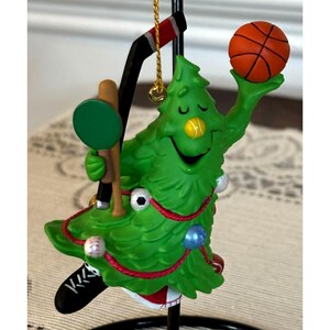 May include: A whimsical Christmas tree ornament featuring a smiling green tree holding a basketball. The tree also has a hockey stick, baseball bat, and other sports-themed decorations. The ornament is suspended from a gold-colored string.