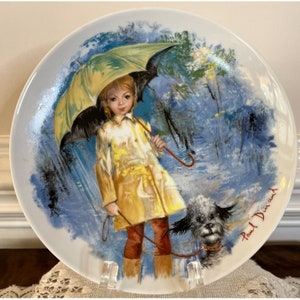 May include: A vintage porcelain plate featuring a painting of a girl in a yellow raincoat walking a dog in the rain. The girl is holding a yellow and black umbrella. The plate is signed "Paul Durand".
