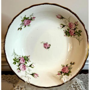 May include: A white ceramic serving bowl with a scalloped edge and a gold rim. The bowl is decorated with pink roses and green leaves. The bowl is displayed on a clear stand.