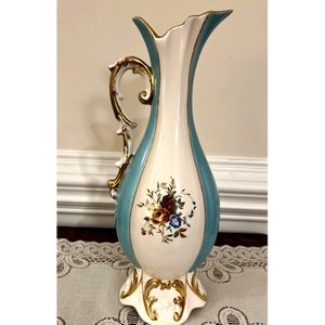 May include: A white and blue porcelain vase with a gold handle and floral design. The vase has a flared top and a round base.