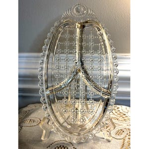 May include: A clear, oval-shaped glass serving dish with three compartments. The dish has a beaded edge and a decorative pattern of small circles. It is supported by a clear plastic stand, and is displayed on a lace tablecloth.