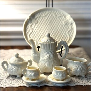 May include: A white porcelain tea set with a delicate floral design. The set includes a teapot, creamer, sugar bowl, two teacups, and two saucers. The set is decorated with a woven pattern and floral accents.