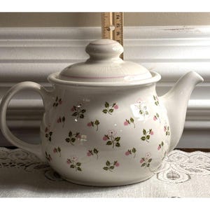 May include: Cream-colored ceramic teapot with a lid, handle, and spout. The teapot is decorated with small pink flowers and green leaves. A ruler is placed behind the teapot. The teapot is approximately 6 inches tall.