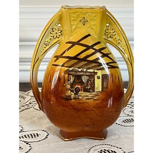 May include: A yellow and brown ceramic vase with two handles. The vase is decorated with a painted scene of a fireplace and people inside a house.