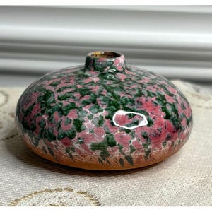 May include: A small, round ceramic vase with a wide, flattened body and a narrow neck. The surface features a mottled, speckled glaze pattern in shades of deep forest green and dusty rose pink, with a smooth, glossy finish over the upper portion.