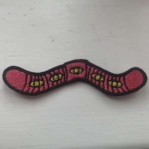 Puede incluir: Un parche bordado de gusano de color rosa y amarillo con costuras negras.