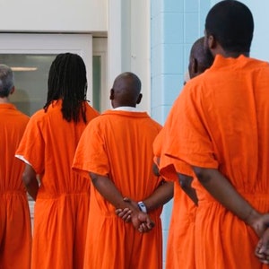 May include: Five people wearing orange jumpsuits stand in a line, facing away from the camera. They have their hands clasped behind their backs.