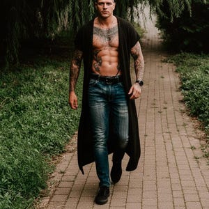 Könnte beinhalten: Eine Person in einem schwarzen Cardigan mit offenem Schnitt, blauen Jeans und schwarzen Schuhen geht auf einem Ziegelweg. Die Person hat Tätowierungen auf Armen und Brust. Eine schwarze Uhr befindet sich an ihrem linken Handgelenk.