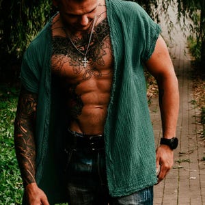 Peut inclure: Une chemise vert foncé à devant ouvert et à manches courtes. La chemise est faite d'un tissu texturé. La personne porte un collier en argent avec un pendentif en forme de croix et une montre noire. La personne porte également un jean bleu.