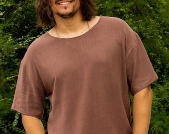 Könnte beinhalten: Ein braunes Kurzarm-Shirt mit lockerer Passform. Das Shirt hat einen Rundhalsausschnitt und ist aus einem weichen, strukturierten Stoff gefertigt. Die Person trägt Sonnenbrille und Armbänder. Der Hintergrund ist üppiges grünes Laub.