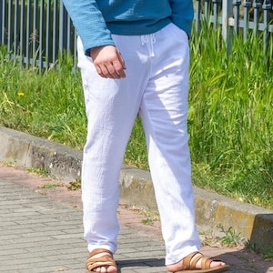 May include: White linen pants with a drawstring waist. The pants are paired with brown leather sandals. The person is wearing a blue long-sleeve shirt. The pants appear to be made of a lightweight, textured fabric.
