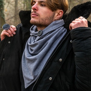 May include: A grey scarf with a button fastening. The scarf is being worn by a person wearing a black jacket.