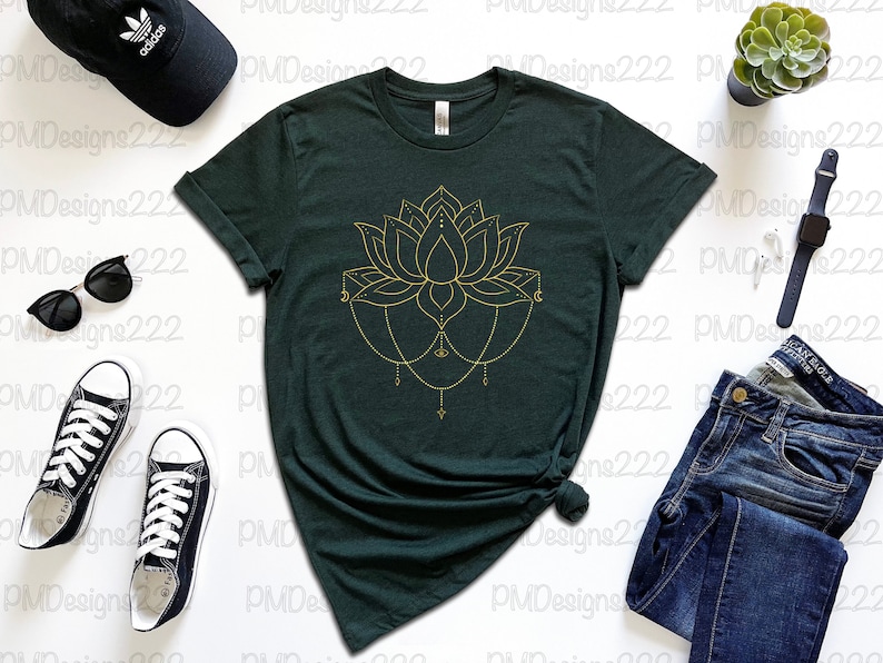 Puede incluir: Camiseta verde oscuro con un dise&ntilde;o de flor de loto dorada. La imagen incluye vaqueros azules, gafas de sol negras, una gorra negra y zapatillas deportivas blancas y negras. Tambi&eacute;n se ven una peque&ntilde;a planta suculenta y un reloj inteligente.