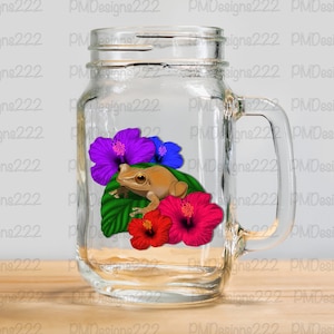 May include: A clear glass mason jar mug with a handle. Inside the mug is a brown frog sitting on a green leaf with red, purple, and blue hibiscus flowers.