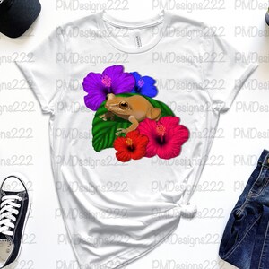 May include: A white t-shirt featuring a brown frog perched on a green leaf with pink, blue and purple hibiscus flowers.