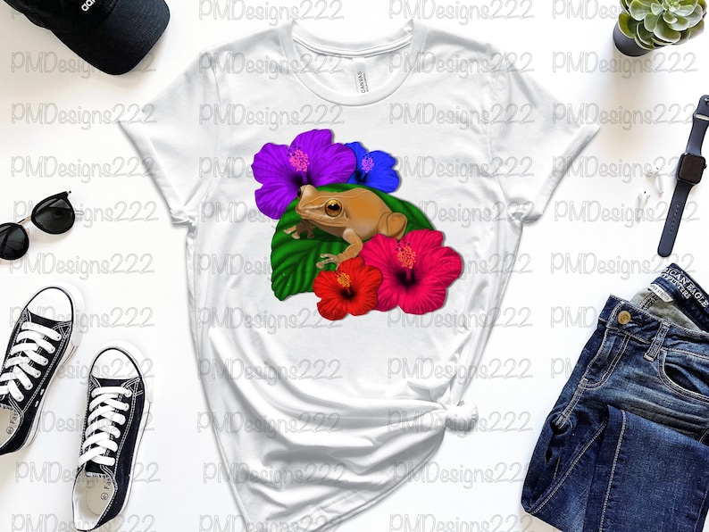 May include: A white t-shirt featuring a brown frog perched on a green leaf with pink, blue and purple hibiscus flowers.