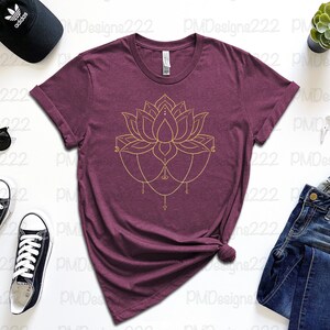 Puede incluir: Una camiseta granate con un dise&ntilde;o de flor de loto dorada. La imagen incluye vaqueros azules, gafas de sol negras, una gorra de b&eacute;isbol negra y zapatillas negras. Tambi&eacute;n se ven una planta suculenta y un reloj inteligente.