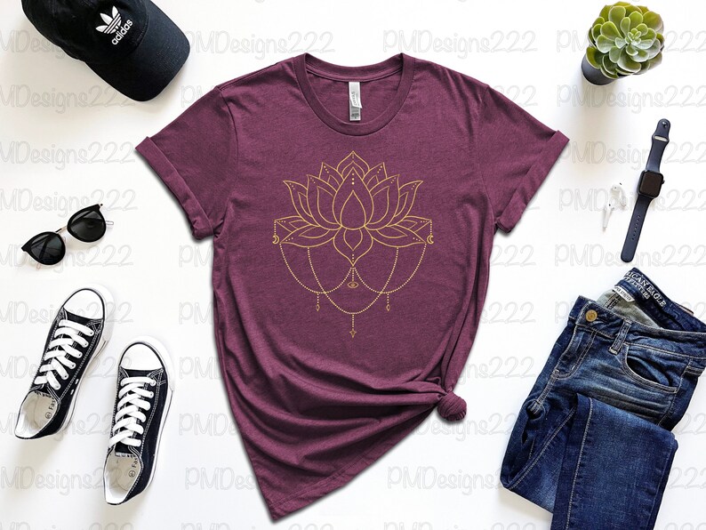 Puede incluir: Una camiseta granate con un dise&ntilde;o de flor de loto dorada. La imagen incluye vaqueros azules, gafas de sol negras, una gorra de b&eacute;isbol negra y zapatillas negras. Tambi&eacute;n se ven una planta suculenta y un reloj inteligente.