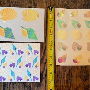 May include: Three sheets of seashell stickers featuring various holographic and colorful designs. A yellow measuring tape is placed vertically between the sheets, showing a length of approximately 10 inches for the tape measure.