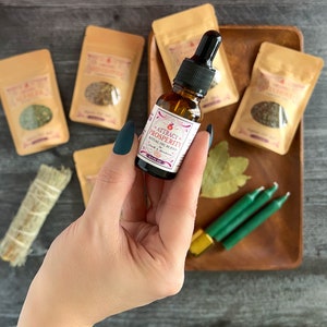 May include: A small glass bottle with a dropper filled with a clear liquid. The bottle has a white label with the text "ATTRACT PROSPERITY RITUAL OIL BLEND" in pink and black lettering. The bottle is surrounded by small brown paper bags, green candles, and a bay leaf on a wooden tray.