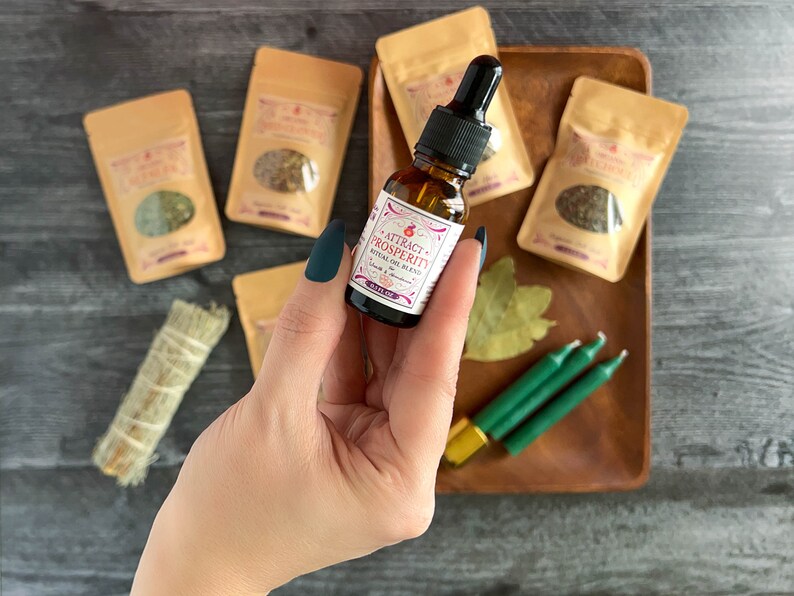 May include: A small glass bottle with a dropper filled with a clear liquid. The bottle has a white label with the text "ATTRACT PROSPERITY RITUAL OIL BLEND" in pink and black lettering. The bottle is surrounded by small brown paper bags, green candles, and a bay leaf on a wooden tray.