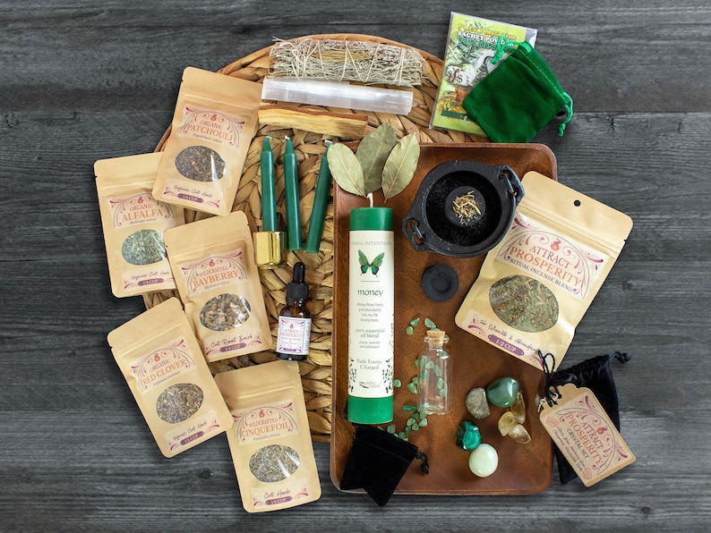 May include: A collection of green and brown herbal incense blends, green candles, a green candle with a butterfly design and the word "money", a small black cauldron, a small bottle of oil, a small bag of green crystals, and a small green pouch. The items are arranged on a woven wicker tray with a brown leather tray on top.
