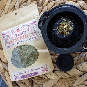 May include: A brown paper bag with a label that reads "Attract Prosperity Ritual Incense Blend" and "For Wealth & Abundance 1/2 Cup". The bag contains dried herbs. A small black cast iron cauldron with a handle is filled with dried herbs. A black charcoal disc is next to the cauldron.