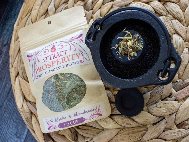 May include: A brown paper bag with a label that reads "Attract Prosperity Ritual Incense Blend" and "For Wealth & Abundance 1/2 Cup". The bag contains dried herbs. A small black cast iron cauldron with a handle is filled with dried herbs. A black charcoal disc is next to the cauldron.