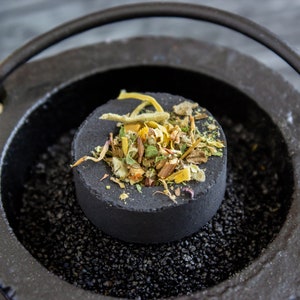 May include: A black cast iron incense burner with a round handle. A black charcoal disc with dried herbs and flowers is placed inside the burner.