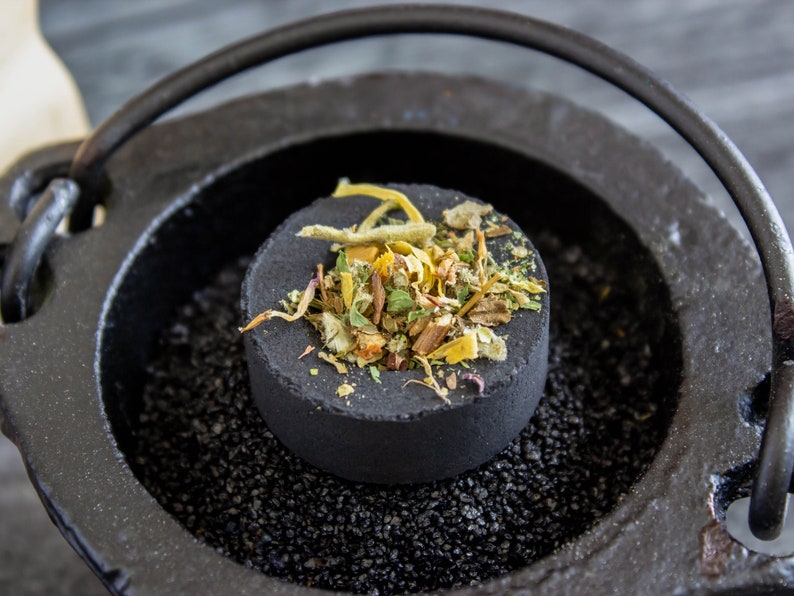 May include: A black cast iron incense burner with a round handle. A black charcoal disc with dried herbs and flowers is placed inside the burner.