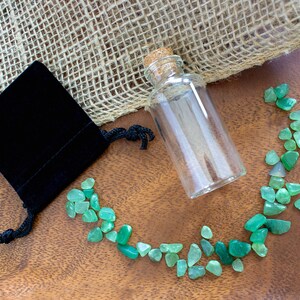May include: A small glass bottle with a cork stopper, a black velvet pouch, and a scattering of green gemstones on a wooden surface.