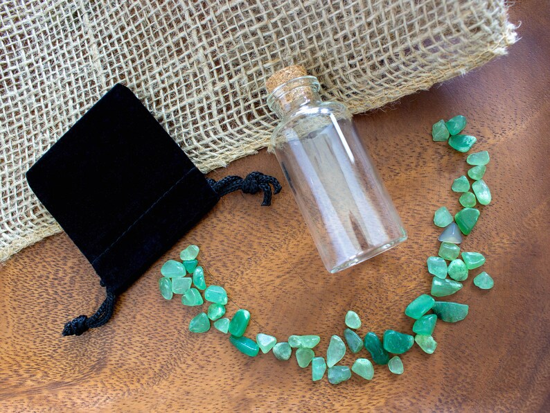 May include: A small glass bottle with a cork stopper, a black velvet pouch, and a scattering of green gemstones on a wooden surface.