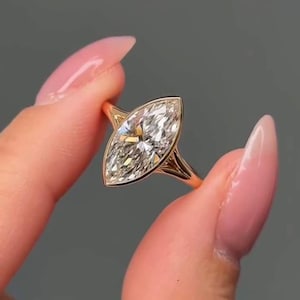 Könnte beinhalten: Ein goldener Ring mit einem großen Diamanten im Marquiseschliff. Der Diamant ist in einer filigranen, offenen Ringschiene gefasst. Der Ring wird vor einem grauen Hintergrund gehalten, um das Funkeln des Edelsteins hervorzuheben.