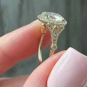 Könnte beinhalten: Ein goldener Verlobungsring mit einem großen, oval geformten, klaren Edelstein. Der Edelstein ist von einem Halo aus kleineren Steinen umgeben. Der Ring hat ein Band und eine Fassung mit aufwendigen Details und zusätzlichen kleinen Steinen.