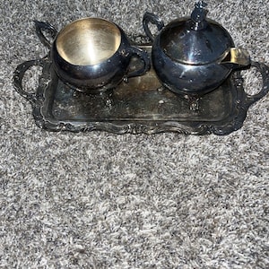 May include: A silver-toned metal tea service set consisting of a rectangular tray with ornate handles, an open sugar bowl with two handles, and a lidded creamer with a single handle and a spout, all featuring a polished metallic finish.